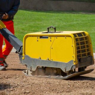 عامل بناء يقوم بتحريك ماكينة رصّ التربة لونها أصفر فوق طريق ترابي جانب مرج أخضر