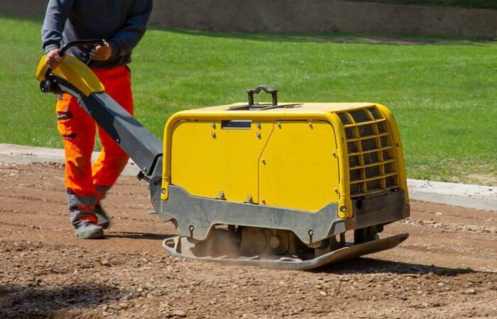 عامل بناء يقوم بتحريك ماكينة رصّ التربة لونها أصفر فوق طريق ترابي جانب مرج أخضر