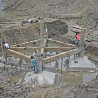 خمسة عمال يقومون بصب أساسات سطحية لأربعة عواميد في حفرة يوجد فيها مياه وجانب الحفرة يوجد غطاس أخضر اللون يقوم بسحب المياه من الحفرة