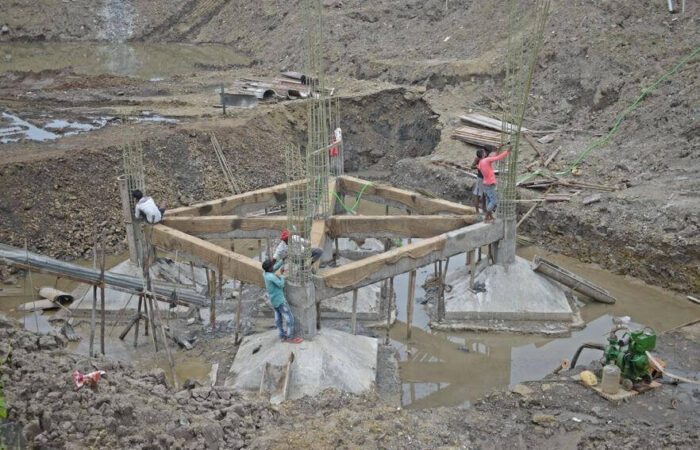 خمسة عمال يقومون بصب أساسات سطحية لأربعة عواميد في حفرة يوجد فيها مياه وجانب الحفرة يوجد غطاس أخضر اللون يقوم بسحب المياه من الحفرة