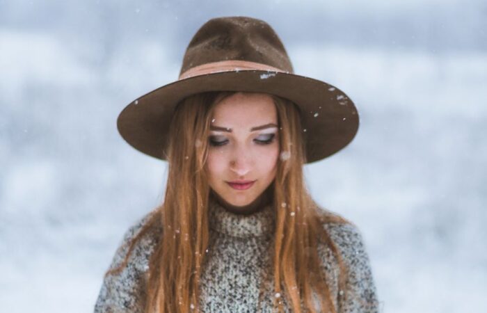 فتاة واقفة في الخارج وهي تلبس كنزة صوف رمادية وتضع قبعة بنية على رأسها وتنظر للأسفل بحزن ويظهر الثلج من خلفها