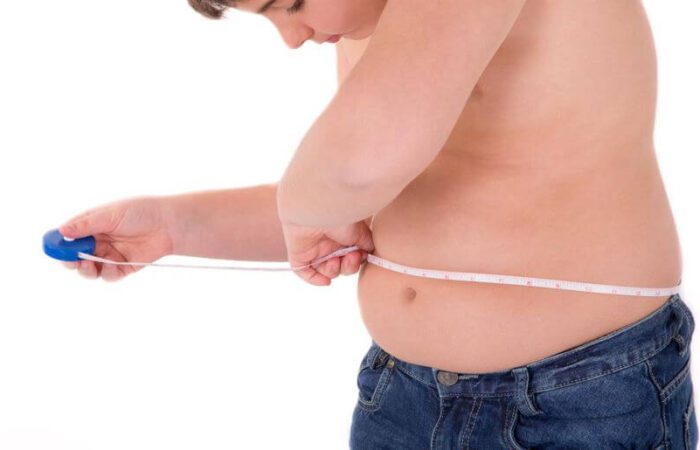 طفل سمين يظهر نصف عارياً يلبس بنطلون جينز أزرق اللون ويلف شريط قياس حول بطنه وهو ينظر للرقم الظاهر على شريط القياس