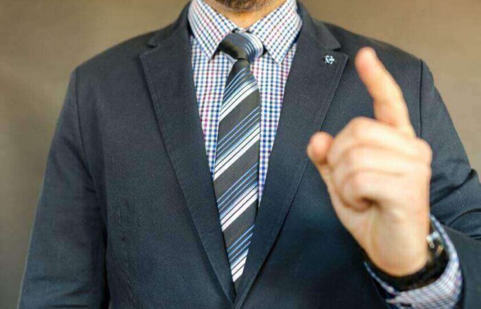 شخص بالغ يرتدي بدلة رسمية لونها رمادي وتحتها قميص منقط ويقوم بالإشارة بسبابة يده نحو الأعلى