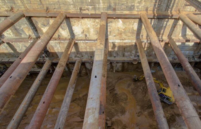 مواسير معدنية ضحمة ممتدة بشكل أفقي تقوم بتدعيم جوانب حفرة لمنشئة هندسية ضخمة ويوجد داخل الحفرة حفار أصفر يقوم بأعمال الحفر