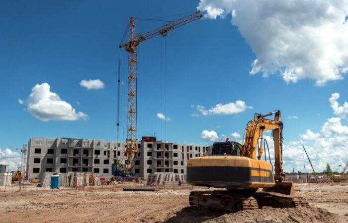 حفار آلي يقوم بجرف التربة أمام بناية سكنية قيد الإنشاء ويوجد أمام البناء رافعة صفراء طويلة