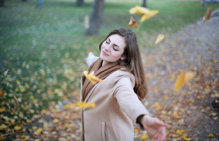 فتاة بالغة وسط حديقة وهي تلبس معطفا بني اللون تقوم بفتح زراعيها سعيدة بتساقط أوراق الشجر