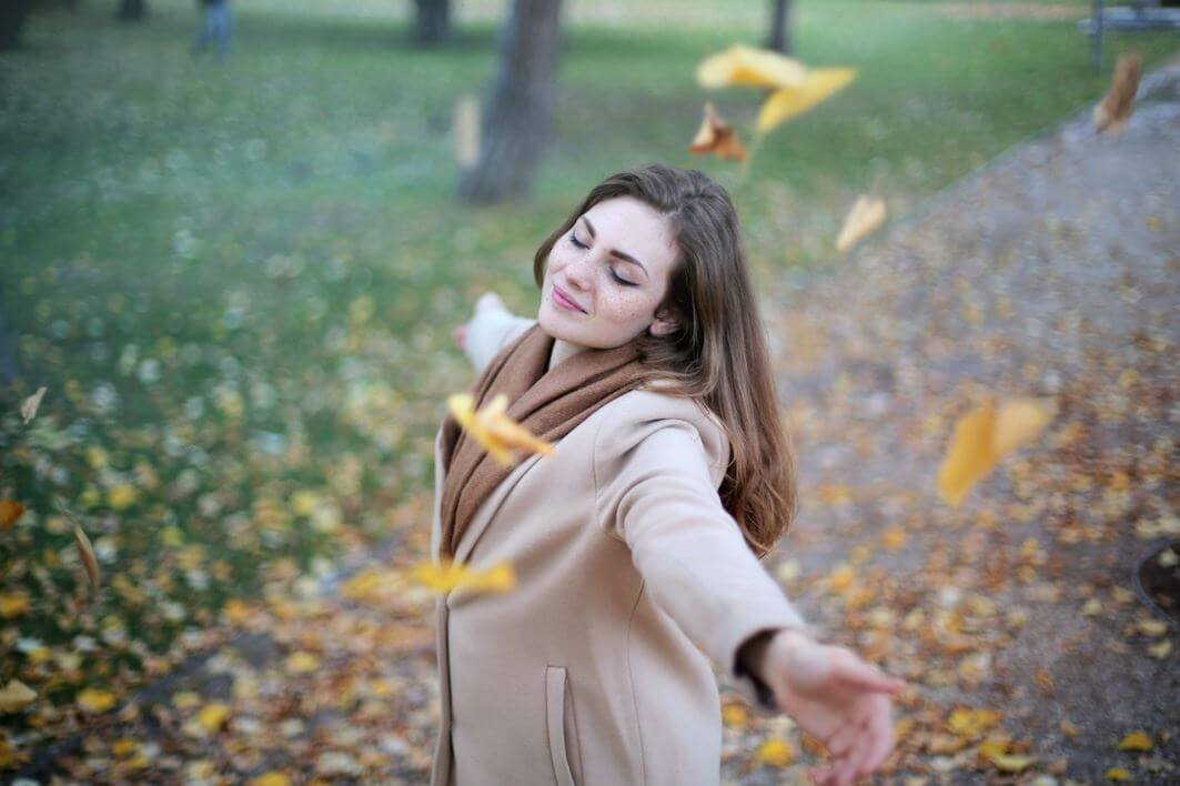 فتاة بالغة وسط حديقة وهي تلبس معطفا بني اللون تقوم بفتح زراعيها سعيدة بتساقط أوراق الشجر