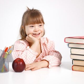 فتاة من ذوي الاحتياجات الخاصة تجلس مبتسمة أمام طاولة يوجد عليها كتب و تفاحة بالإضافة الى أقلام وتقوم الفتاة بوضع يدها أسفل رأسها