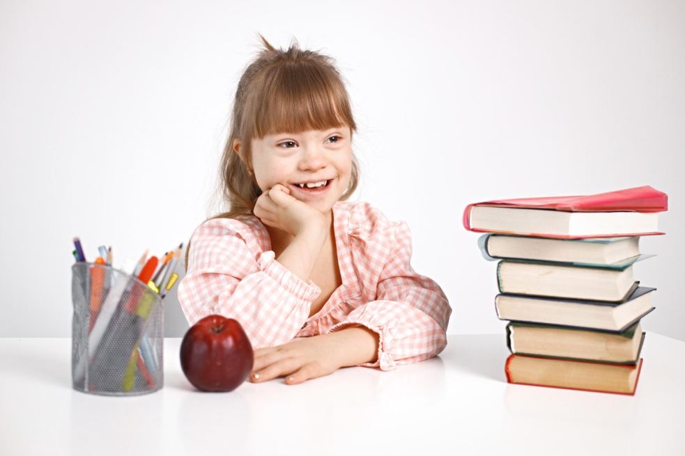 فتاة من ذوي الاحتياجات الخاصة تجلس مبتسمة أمام طاولة يوجد عليها كتب و تفاحة بالإضافة الى أقلام وتقوم الفتاة بوضع يدها أسفل رأسها