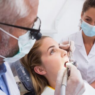 طبيب أسنان يقوم بإدخال أداة الحفر في فم المريضة وتوجد مساعدة دكتور الأسنان وهي جالسة بجانب المريضة وتراقبها، ويلبس كل من الدكتور والمساعدة كمامات واقية