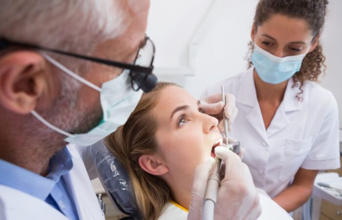 طبيب أسنان يقوم بإدخال أداة الحفر في فم المريضة وتوجد مساعدة دكتور الأسنان وهي جالسة بجانب المريضة وتراقبها، ويلبس كل من الدكتور والمساعدة كمامات واقية