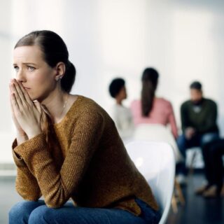 فتاة بالغة تلبس كنزة بنية اللون وتجلس وحيدة بعيداً عن مجموعة من الناس خلفها وتضع يديها بالقرب من فمها وهي تبدو خائفة من الحالة التي هي فيها
