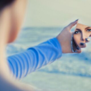 امرأة بالغة ترتدي كنزة زرقاء وتنظر لنفسها في مرآة مكسورة على شاطئ البحر