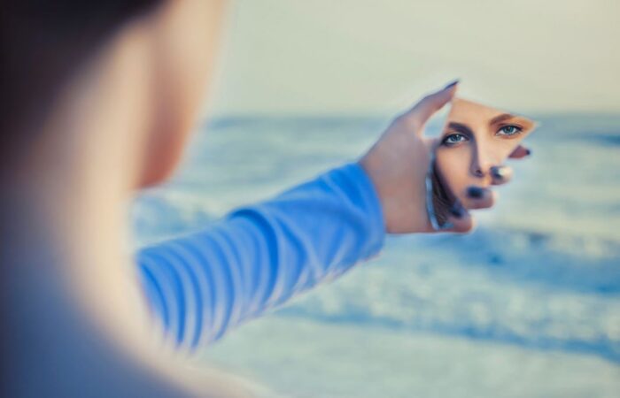 امرأة بالغة ترتدي كنزة زرقاء وتنظر لنفسها في مرآة مكسورة على شاطئ البحر