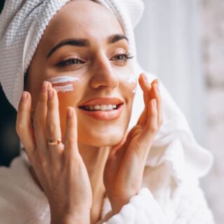 فتاة بالغة تضع منشفة بيضاء على رأسها وتقوم بوضع كريم أبيض على خدها وهي تبتسم