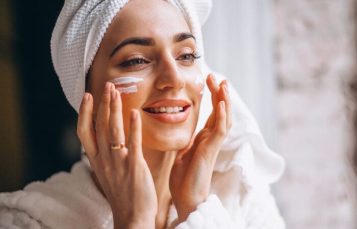 فتاة بالغة تضع منشفة بيضاء على رأسها وتقوم بوضع كريم أبيض على خدها وهي تبتسم