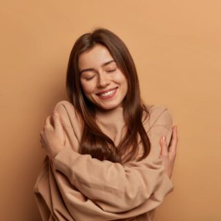 امرأة بالغة تقوم بضم نفسها من خلال لف يديها حول نفسها وهي مغمضة العينين وتبتسم وتظهر عليها علامات الرضى النفسي