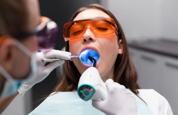 طبيبة أسنان تقوم بمعالجة سن مريضة عن طريق شعاع ليزر أزرق اللون ويلبس كل من الطبيبة والمريضة نظارات واقية من أشعة الليزر لونها أورانج
