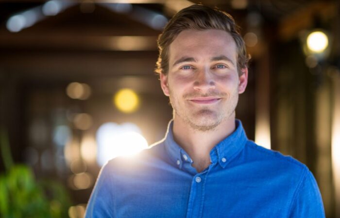 رجل بالغ يلبس قميص أزرق اللون و يقف مبتسما في الحديقة مساءً وتظهر على وجهه علامات الإعجاب