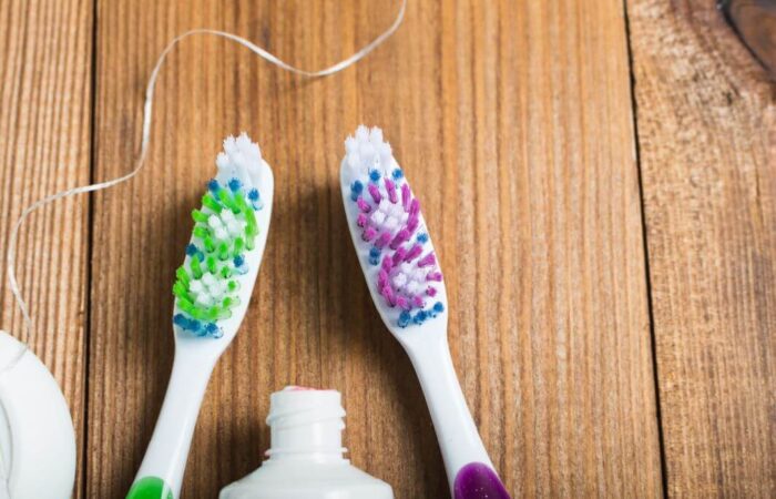 فرشاتين أسنان على لوح خشبي وبينهما معجون أسنان وعلى الجانب توجد علبة يخرج منها خيط لتنظيف الأسنان