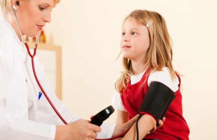 فتاة صغيرة تلبس ثوب أحمر وتجلس في عيادة الطبيب ويقوم الطبيب بلف جهاز قياس الضغط حول يدها اليسار لقياس ضغطها والاستماع الى نبضات قلبها