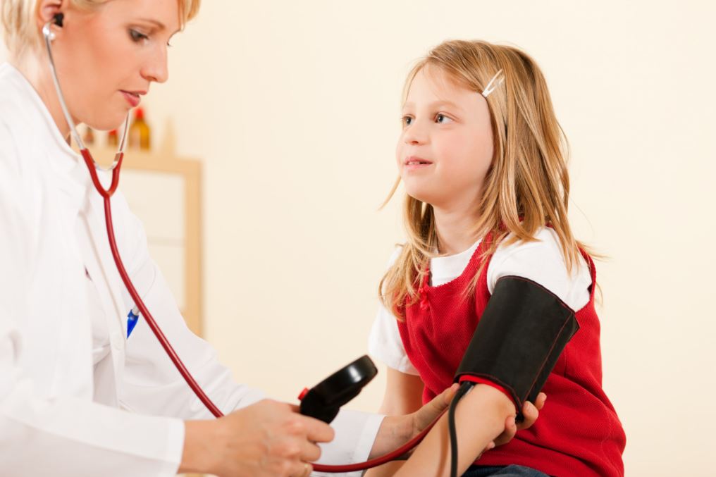 فتاة صغيرة تلبس ثوب أحمر وتجلس في عيادة الطبيب ويقوم الطبيب بلف جهاز قياس الضغط حول يدها اليسار لقياس ضغطها والاستماع الى نبضات قلبها