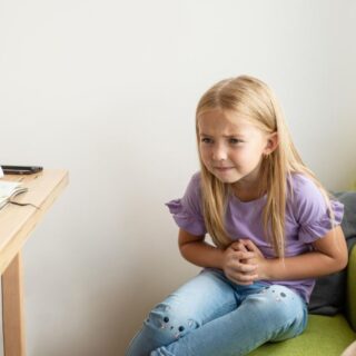 فتاة شقراء صغيرة جالسة في غرفتها أمام طاولة الدراسة وتضع يديها على بطنها وتضغط عليه بقوة بسبب احساسها بألم في منطقة البطن