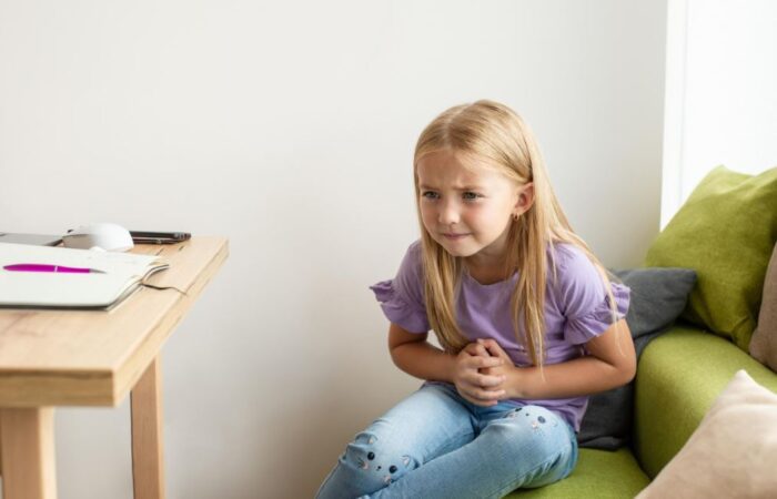 فتاة شقراء صغيرة جالسة في غرفتها أمام طاولة الدراسة وتضع يديها على بطنها وتضغط عليه بقوة بسبب احساسها بألم في منطقة البطن