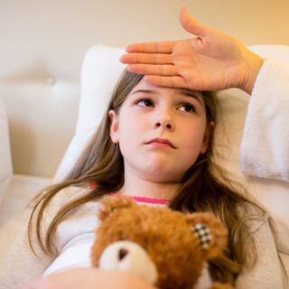 فتاة صغيرة ممددة في سريرها تقوم بحضن لعبة الدب وتظهر عليها علامات المرض وتضع والدتها يدها على جبين الفتاة