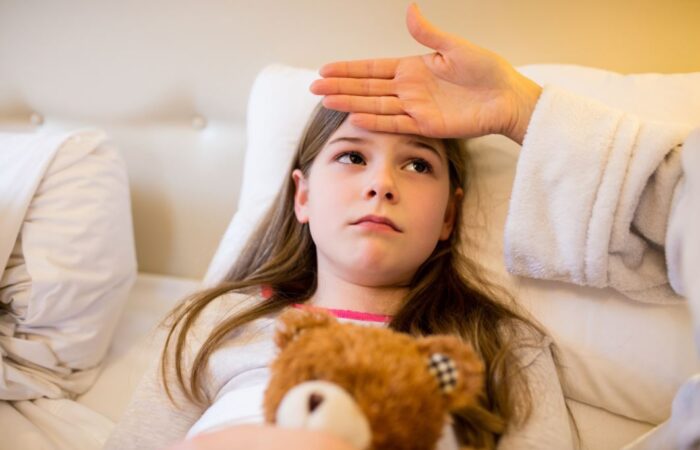 فتاة صغيرة ممددة في سريرها تقوم بحضن لعبة الدب وتظهر عليها علامات المرض وتضع والدتها يدها على جبين الفتاة