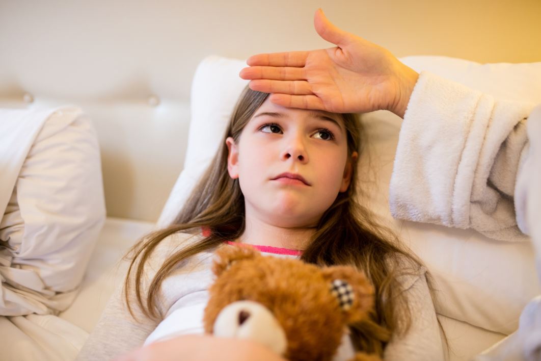 فتاة صغيرة ممددة في سريرها تقوم بحضن لعبة الدب وتظهر عليها علامات المرض وتضع والدتها يدها على جبين الفتاة