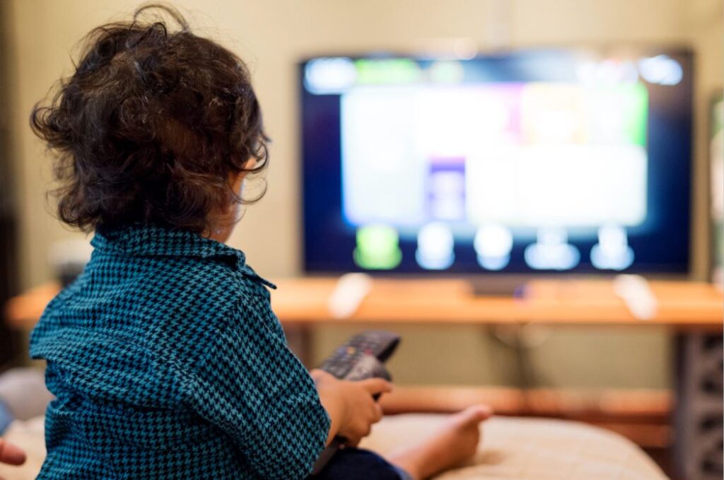 صبي صغير يرتدي قميص أزرق يجلس في غرفة الجلوس أمام التلفاز ويحمل بيده جهاز التحكم