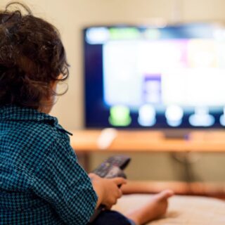 صبي صغير يرتدي قميص أزرق يجلس في غرفة الجلوس أمام التلفاز ويحمل بيده جهاز التحكم