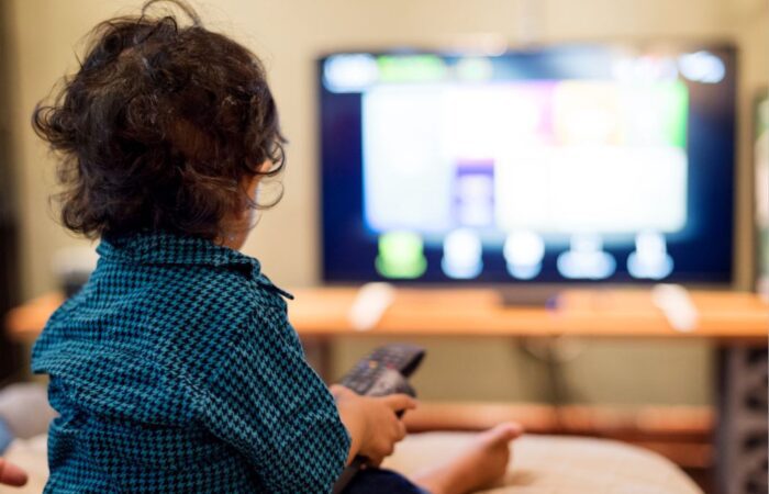 صبي صغير يرتدي قميص أزرق يجلس في غرفة الجلوس أمام التلفاز ويحمل بيده جهاز التحكم