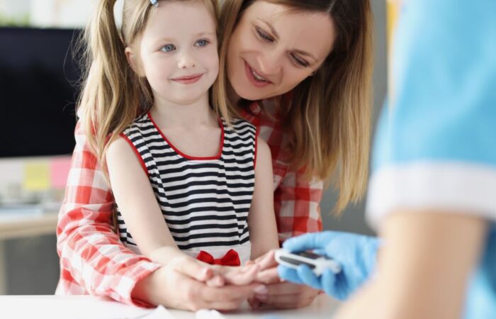 فتاة صغيرة في عيادة الطبيب مع والدتها ويقوم الطبيب بأخذ عينة من دمها لفحص مستوى سكر الدم و ذلك من أجل علاج السكري من النمط الأول لديها وتقوم الأم بحضن الفتاة من الخلف