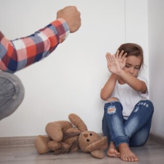 أب يرفع يده على ابنته الصغيرة التي تجلس في زاوية الغرفة وبجانبها لعبة الدب وتقوم الفتاة الصغيرة بوضع يديها أمام وجها لحماية نفسها من والدها