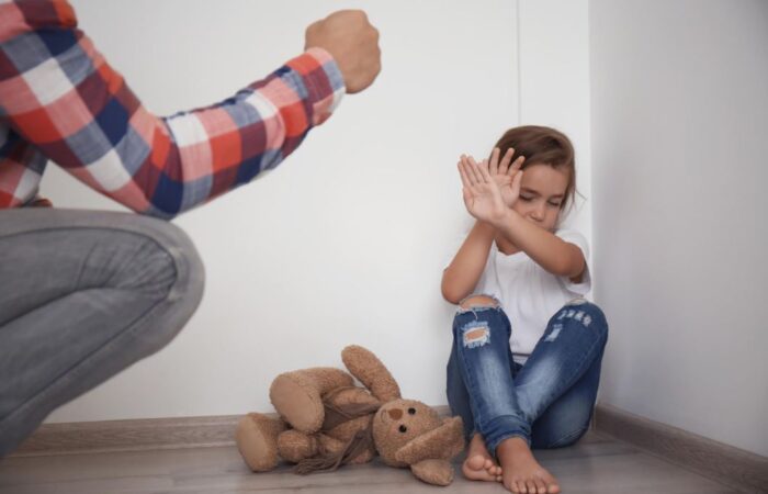 أب يرفع يده على ابنته الصغيرة التي تجلس في زاوية الغرفة وبجانبها لعبة الدب وتقوم الفتاة الصغيرة بوضع يديها أمام وجها لحماية نفسها من والدها