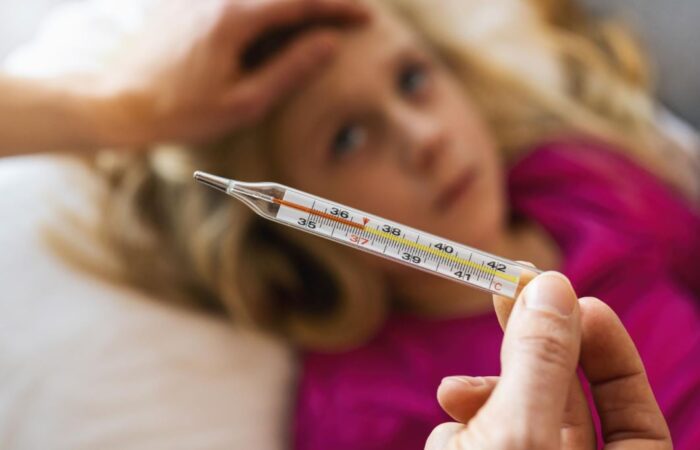 فتاة صغيرة مستلقية في سريرها وتقوم الأم بفحص درجة حرارتها باستخدام ميزان قياس درجة الحرارة وتقوم الأم بوضع يدها الأخرى على جبين ابنتها