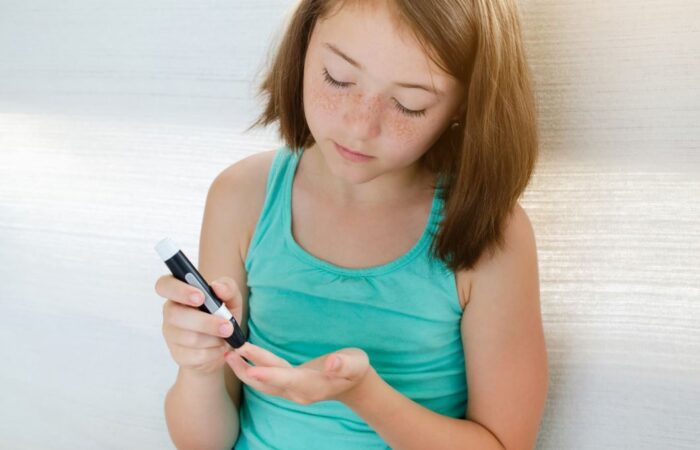 فتاة صغيرة شعرها خمري تلبس كنزة صيفية وتقوم بأخذ عينة من الدم من طرف اصبعها لتفحص مستوى سكر الدم لديها