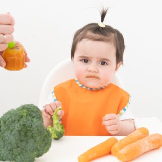 طفلة صغيرة ترتدي كنزة برتقالية اللون تجلس امام طاولة الطعام ويوجد امامها على الطاولة جزر وبروكلي وتقوم الوالدة بإطعامها