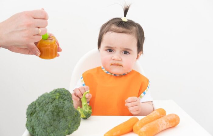 طفلة صغيرة ترتدي كنزة برتقالية اللون تجلس امام طاولة الطعام ويوجد امامها على الطاولة جزر وبروكلي وتقوم الوالدة بإطعامها