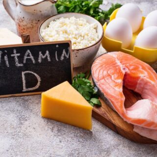 مجموعة من المأكولات مثل فيليه سمك، البيض، الجبنة الصفراء موجودة على طاولة بيضاء و وسطها لافتة مكتوب عليها كلمة فيتامين D بالإنكليزية