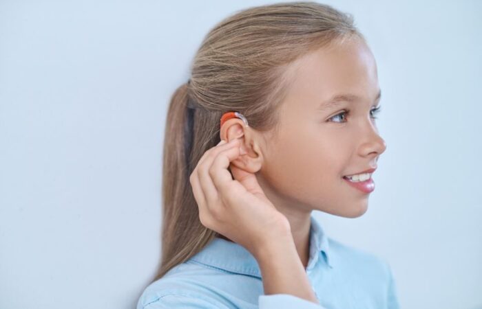 فتاة صغيرة شقراء تلبس قميص أزرق وتقوم بلمس أذنها اليمين التي يوجد عليها جهاز لتقوية السمع