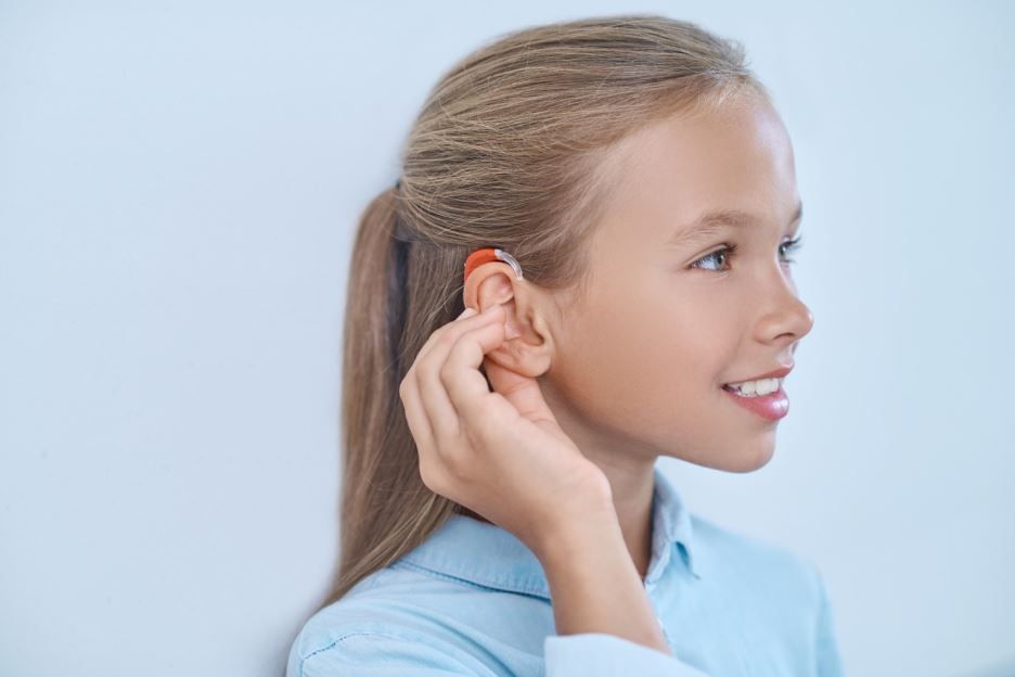 فتاة صغيرة شقراء تلبس قميص أزرق وتقوم بلمس أذنها اليمين التي يوجد عليها جهاز لتقوية السمع
