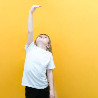 فتاة صغيرة شعرها أشقر تقف أمام حائط لونه أصفر فاتح وترفع يدها فوق رأسها وهي تنظر لها لترى كم أصبح طولها