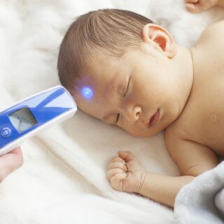 طفل رضيع ينام على سريره وتقوم الأم بفحص درجة حر ارة جسمه من خلال توجيه جهاز قياس درجة حرارة الجسم نحو جبينه