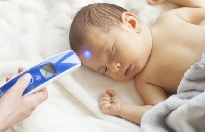 طفل رضيع ينام على سريره وتقوم الأم بفحص درجة حر ارة جسمه من خلال توجيه جهاز قياس درجة حرارة الجسم نحو جبينه