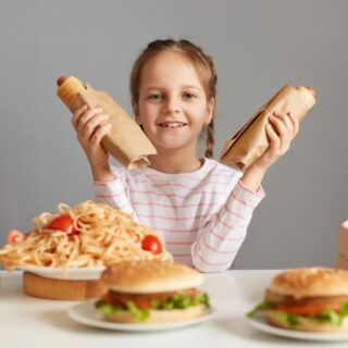 فتاة صغيرة تجلس أمام طاولة مليئة بالوجبات السريعة وتمسك بيديها قطعتين من السندويش