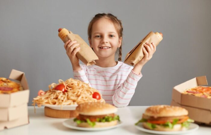 فتاة صغيرة تجلس أمام طاولة مليئة بالوجبات السريعة وتمسك بيديها قطعتين من السندويش
