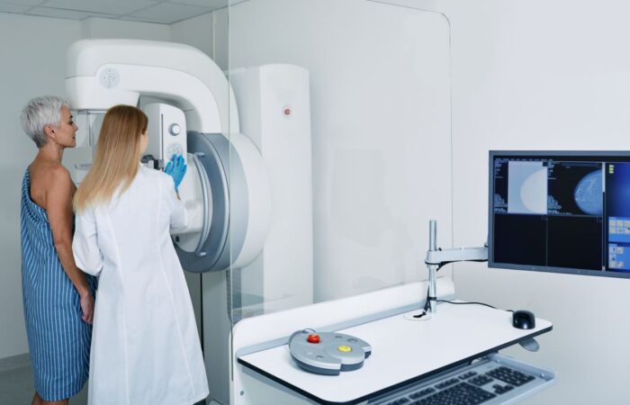 طبيبة نسائية تستخدم جهاز طبي كبير ومن خلفها تقف مريضة مسنّة تلبس ثوب مخطط بالأبيض والأزرق